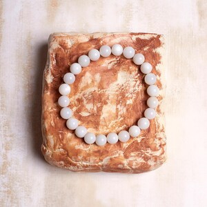 May include: A white beaded bracelet with a round shape, resting on a textured brown surface.