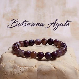 Puede incluir: Un brazalete de &aacute;gata de Botsuana morado con un acabado de piedra natural. El brazalete est&aacute; hecho de cuentas redondas con un patr&oacute;n rayado.