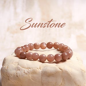 May include: A sunstone bracelet with brown beads. The bracelet is on a white surface. The word "Sunstone" is written in cursive above the bracelet.