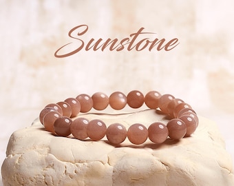 May include: A sunstone bracelet with brown beads. The bracelet is on a white surface. The word "Sunstone" is written in cursive above the bracelet.