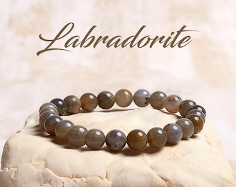 May include: A gray labradorite gemstone bracelet with round beads. The bracelet is on a white surface. Labradorite.