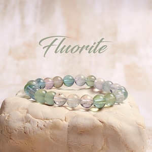 May include: A multi-colored fluorite bracelet with green, blue, and purple beads. The bracelet is on a white rock with the word "Fluorite" written above it.
