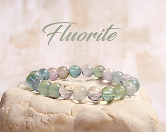 May include: A multi-colored fluorite bracelet with green, blue, and purple beads. The bracelet is on a white rock with the word "Fluorite" written above it.