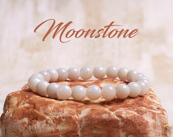 May include: A white moonstone bracelet with round beads. The bracelet is resting on a brown rock.