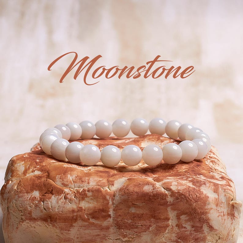 May include: A white moonstone bracelet with round beads. The bracelet is resting on a brown rock.