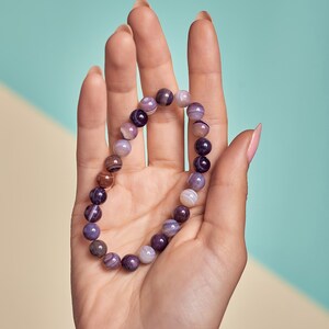 Puede incluir: Un brazalete de cuentas de &aacute;gata de color morado, marr&oacute;n y blanco. El brazalete est&aacute; hecho de cuentas redondas con un acabado de piedra natural.
