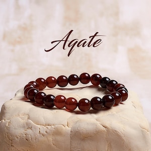 May include: A brown agate gemstone bracelet with round beads. The bracelet is on a white surface. Agate is written in brown text above the bracelet.