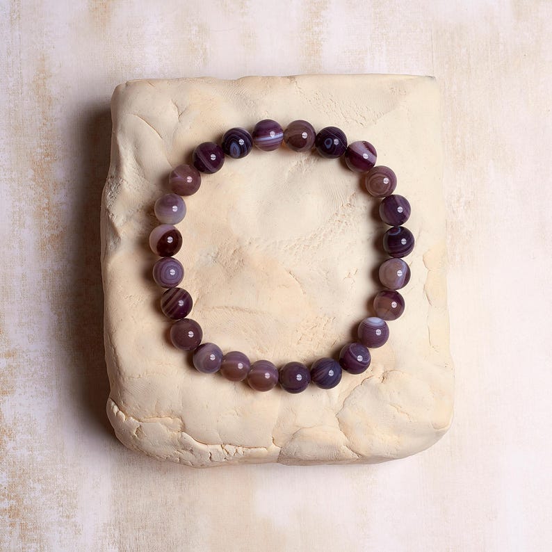 Puede incluir: Un brazalete de cuentas de &aacute;gata p&uacute;rpura con una banda el&aacute;stica extensible. Las cuentas son redondas y tienen un acabado natural y pulido.