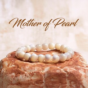 May include: A white mother of pearl bracelet with iridescent beads. The bracelet is on a brown textured surface.