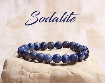 May include: A blue sodalite gemstone bracelet with round beads. The bracelet is on a white stone surface. The word "Sodalite" is written in cursive above the bracelet.