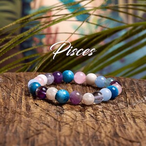 May include: A beaded bracelet with various colored gemstones, including blue, purple, pink, and white, is displayed on a wooden surface. The word "Pisces" is written in cursive script above the bracelet. The background features green foliage.