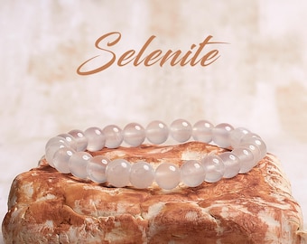 May include: A white selenite beaded bracelet on a brown textured surface. The bracelet is made of smooth, round beads.