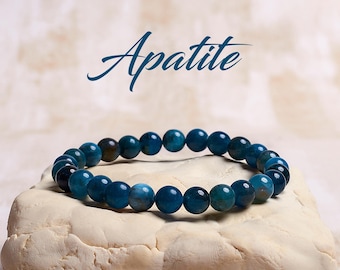 May include: A blue apatite gemstone bracelet with round beads. The bracelet is on a light beige surface. The word "Apatite" is written in blue above the bracelet.