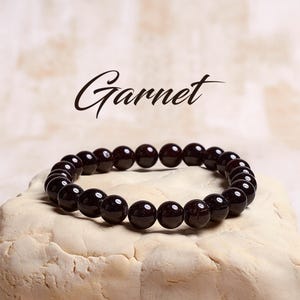May include: A garnet gemstone bracelet with dark red beads. The bracelet is on a light beige surface. Garnet