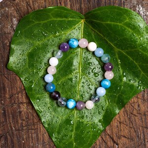 May include: A beaded bracelet composed of various colorful gemstones, including blue, purple, pink, and white, rests on a large, wet green leaf. The bracelet's beads are round and smooth, creating a vibrant and natural aesthetic.
