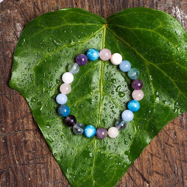 May include: A beaded bracelet composed of various colorful gemstones, including blue, purple, pink, and white, rests on a large, wet green leaf. The bracelet's beads are round and smooth, creating a vibrant and natural aesthetic.