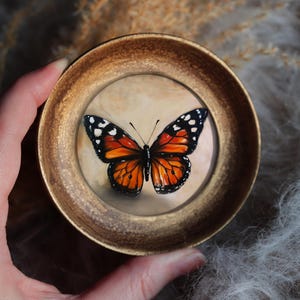 May include: A round, gold-toned frame holds a detailed painting of a butterfly. The butterfly has orange and black wings with white spots. The background is a soft, neutral color. The frame is held by a hand.