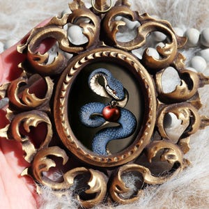 Puede incluir: Un adorno de pared ovalado con un marco de metal dorado ornamentado con volutas intrincadas. El centro muestra una ilustración oscura de una serpiente azul que sostiene una manzana roja entre sus anillos. Tiene una pequeña anilla metálica en la parte superior.