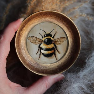May include: A small, round, framed painting of a bee. The bee is depicted with black and yellow stripes, wings, and antennae. The frame is gold-colored. The painting is held in a hand.