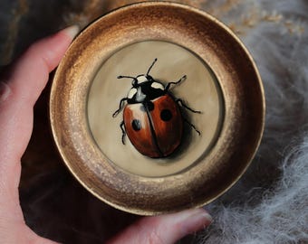 May include: A small, round, gold-colored dish holds a detailed painting of a ladybug. The ladybug is red with black spots and a black head. The dish is held by a hand, and the background is a soft, textured gray.