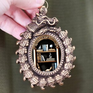 Może przedstawiać: Brązowy wisiorek z motywem smoka na górze i szczegółową owalną ramką. Wewnątrz ramki znajduje się miniaturowa scena biblioteczna z półkami na książki, książkami i globusem. Wisiorek jest trzymany przez dłoń.