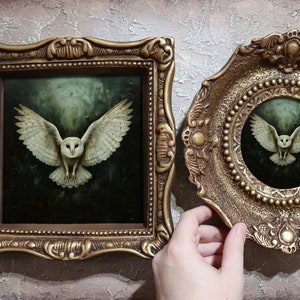 Può includere: Un dipinto quadrato e un piatto decorativo circolare, entrambi raffiguranti un barbagianni bianco in volo su sfondo scuro. Ogni pezzo è racchiuso in una cornice decorata color oro antico con dettagli in rilievo e bordi strutturati.