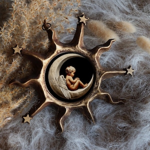 Pode incluir: Uma decoração de parede em forma de sol, cor bronze, com um centro circular apresentando uma imagem de um querubim sentado em uma lua crescente. O sol tem raios pontiagudos e detalhes de estrelas. O fundo é cinza macio e texturizado.