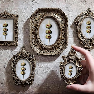 Può includere: Cinque cornici dorate ornate con sfondo bianco e un dipinto di olive verdi su uno spiedino. Le olive hanno centri rossi.