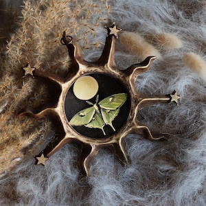 Pode incluir: Um ornamento decorativo em forma de explosão solar com um círculo preto central com uma mariposa verde e uma lua amarela. A explosão solar tem detalhes de estrelas e um acabamento bronze. O fundo é cinza macio e texturizado.