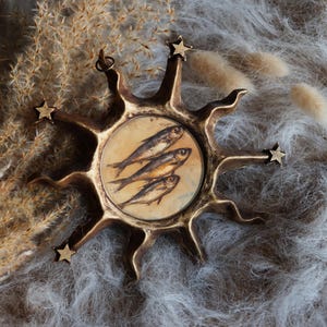 May include: A circular bronze-toned wall hanging featuring a sunburst design with radiating points and small stars at the tips. The centre contains a circular illustration of three fish stacked vertically against a light beige background.