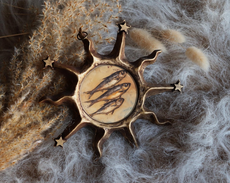 May include: A circular bronze-toned wall hanging featuring a sunburst design with radiating points and small stars at the tips. The centre contains a circular illustration of three fish stacked vertically against a light beige background.