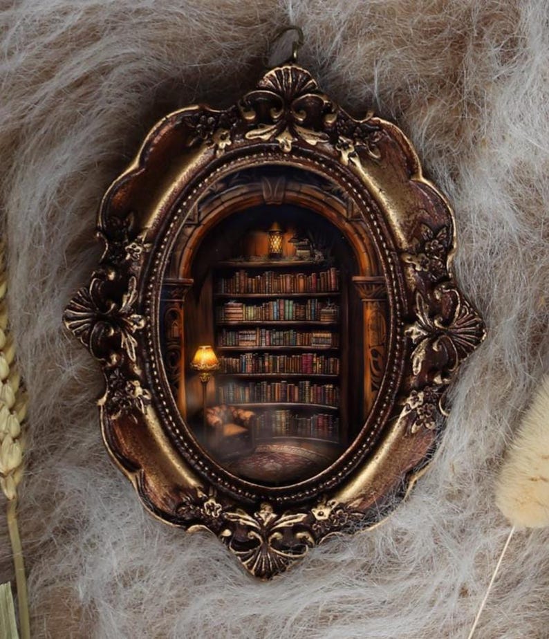 May include: An oval-shaped, ornate, bronze-colored frame with a miniature library scene inside. The library features bookshelves filled with books, a reading lamp, and a cozy armchair. The frame is set against a soft, textured background.
