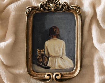 Op de afbeelding: Een ingelijst schilderij van een vrouw in een witte jurk die met een kat zit. De lijst is versierd en goudkleurig.