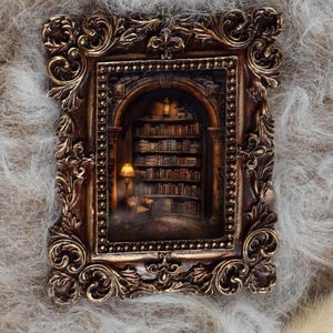 May include: A detailed, ornate brown picture frame with gold accents. Inside, a miniature library scene is illuminated by a warm lamp, with bookshelves and a person reading. The frame is set against a soft, textured background.