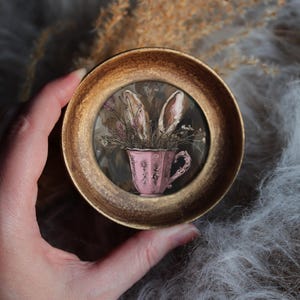 Op de afbeelding: Een klein, rond kunstwerk in een goudkleurige lijst. Het toont een roze theekopje gevuld met gedroogde bloemen en konijnenoren. Het kopje heeft een decoratief handvat en een bloemenpatroon. De achtergrond is zacht en neutraal van kleur.