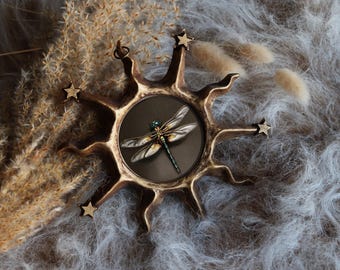 Può includere: Una decorazione da parete circolare con l'immagine centrale di una libellula dalle ali iridescenti e il corpo verde, racchiusa in una cornice color bronzo a forma di sole con raggi appuntiti e piccole stelle sulle punte, dotata di gancio.