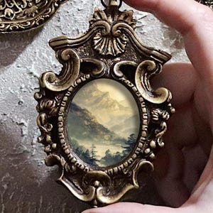 K&ouml;nnte beinhalten: Verziertes Anh&auml;nger im antiken Stil mit einer Miniatur-Landschaftsmalerei. Der Anh&auml;nger hat einen detaillierten, goldfarbenen Rahmen mit Schn&ouml;rkeln und eine kleine Schlaufe zum Aufh&auml;ngen. Das Gem&auml;lde zeigt eine neblige Berglandschaft mit B&auml;umen und einem See.