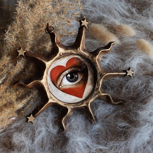 Może przedstawiać: Złoty wisiorek w kształcie słońca z namalowanym wzorem oka i serca. Oko jest niebieskie, a serce czerwone. Wisiorek jest otoczony pięcioma małymi złotymi gwiazdami.