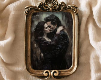 May include: Ornate gold-colored framed artwork featuring a romantic scene of a couple embracing. The artwork depicts a man and woman in dark clothing, their faces close together. The frame has decorative scrollwork and a scalloped top.