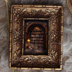 May include: Ornate, gold-colored framed miniature art piece. The artwork depicts a cozy library scene with bookshelves, a reading lamp, and a person. The frame has detailed floral and scrollwork designs, adding to its antique aesthetic.
