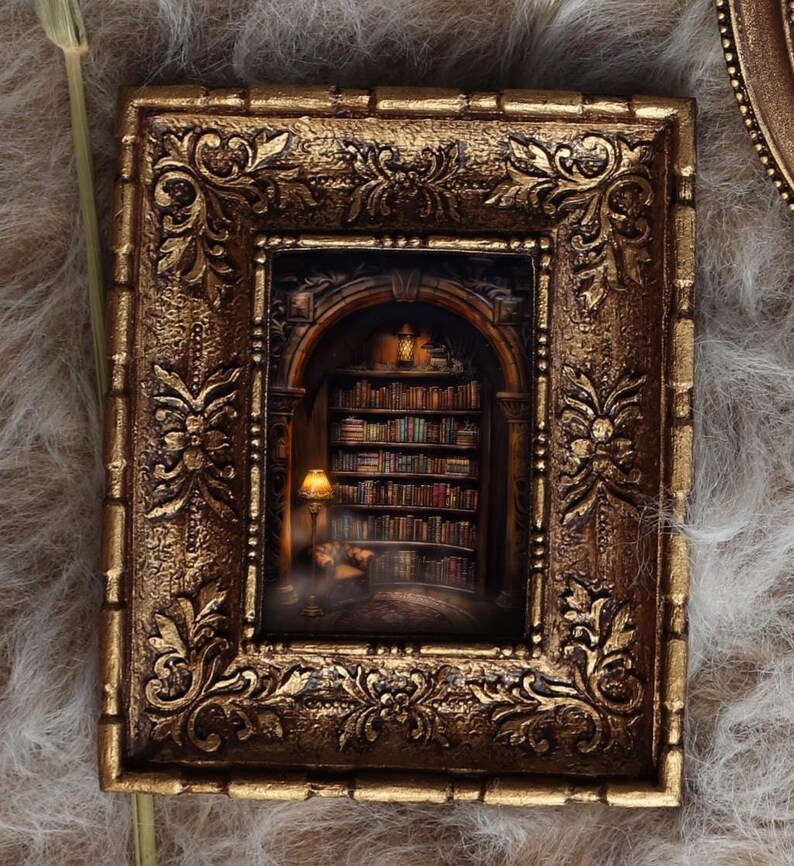 May include: Ornate, gold-colored framed miniature art piece. The artwork depicts a cozy library scene with bookshelves, a reading lamp, and a person. The frame has detailed floral and scrollwork designs, adding to its antique aesthetic.