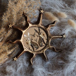 May include: A gold-toned sunburst-shaped ornament with a circular glass panel featuring a hand-painted illustration of two brown rabbits in a field of flowers.