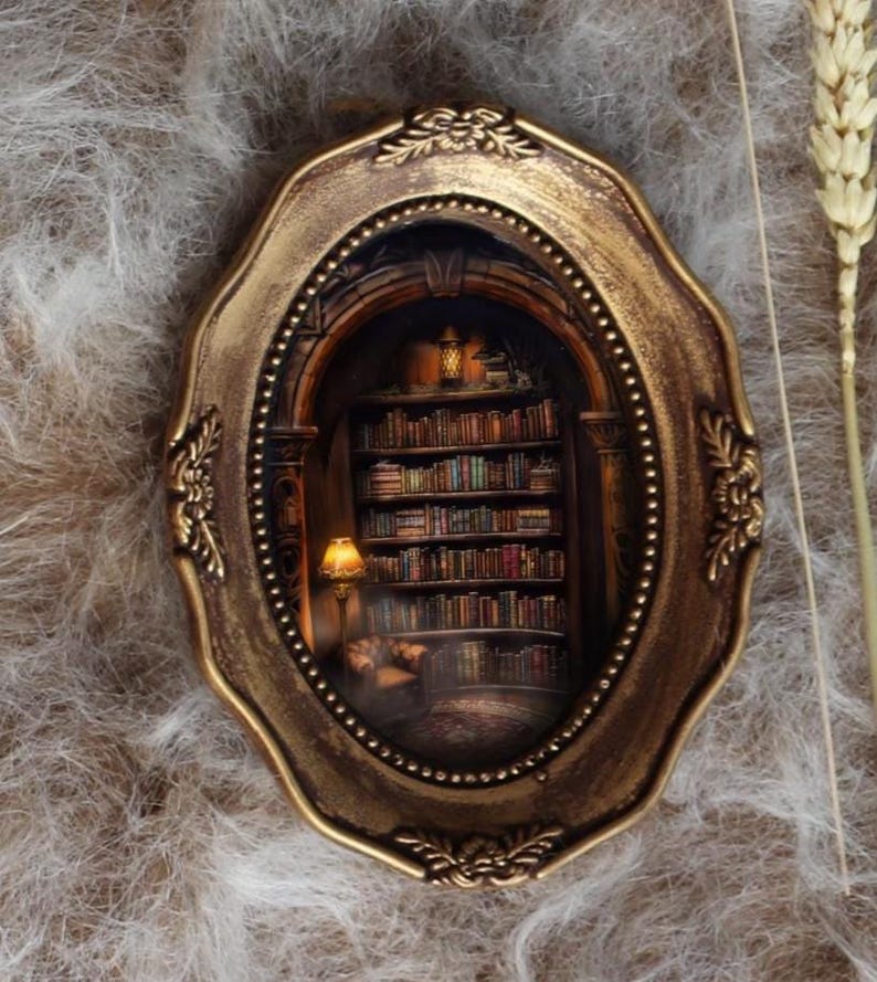 May include: An oval, antique-style frame with a miniature library scene inside. The frame is gold-colored with ornate detailing. The miniature scene features a bookshelf filled with books, a lamp, and a cozy reading nook.