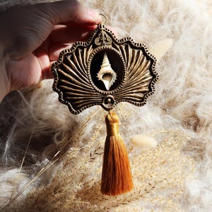 Peut inclure: Pendentif de style antique orné d'un coquillage en son centre. Le cadre de couleur bronze présente un motif détaillé en forme d'éventail. Une gland doré est suspendu en dessous. Le pendentif est tenu sur un fond doux et texturé.