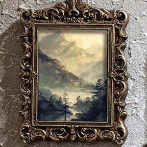 K&ouml;nnte beinhalten: Ein gerahmtes Landschaftsgem&auml;lde mit einer Bergkette, einem Fluss und B&auml;umen. Das Gem&auml;lde befindet sich in einem dekorativen, goldfarbenen Rahmen mit aufwendigen Details. Das Kunstwerk hat eine ged&auml;mpfte Farbpalette mit Gr&uuml;n-, Braun- und Graut&ouml;nen.