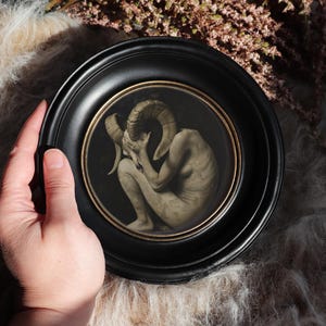 Puede incluir: Un marco circular negro con un borde interior dorado fino que contiene una ilustración de tonos oscuros de una figura pálida con cuernos en posición fetal. La figura tiene grandes cuernos de carnero curvados y se cubre el rostro.