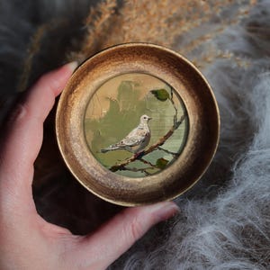 May include: A miniature artwork in a gold-coloured circular frame. The painting depicts a bird on a branch, with a soft colour palette of greens and browns. The artwork has a vintage aesthetic. The frame is approximately 7 cm in diameter.