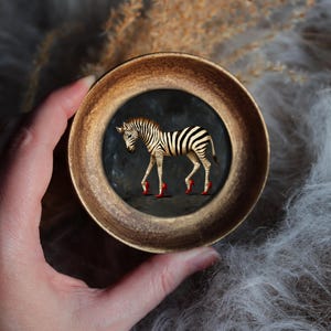 May include: A quirky image of a zebra in red high heels, framed in a circular, gold-coloured mount. The zebra has black and white stripes. The background is dark. The frame is held by a hand.