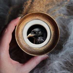 May include: A miniature painting of a black bird's head, set within a round, gold-coloured frame with a white inner border. The bird has a yellow beak and is against a dark backdrop. The frame is held by a hand.