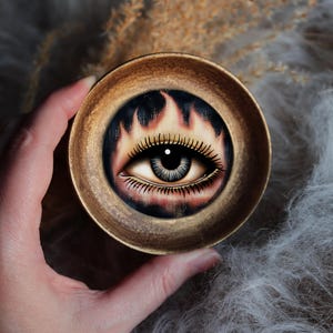 May include: A circular, metallic, bronze-toned bowl featuring a detailed illustration of a human eye centered within a ring of stylized flames. The eye has dark, defined lashes and a realistic iris, set against a dark background within the bowl.